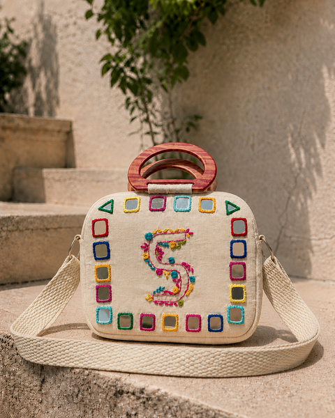 Cream cotton sling bag with hand-embroidered monogram initial and colourful mirrorwork border, wooden top handle and woven crossbody strap