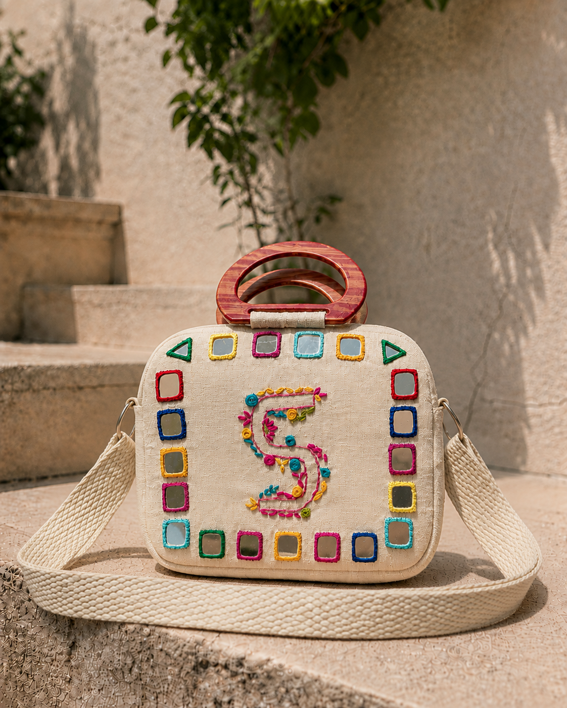 Cream cotton sling bag with hand-embroidered monogram initial and colourful mirrorwork border, wooden top handle and woven crossbody strap 1